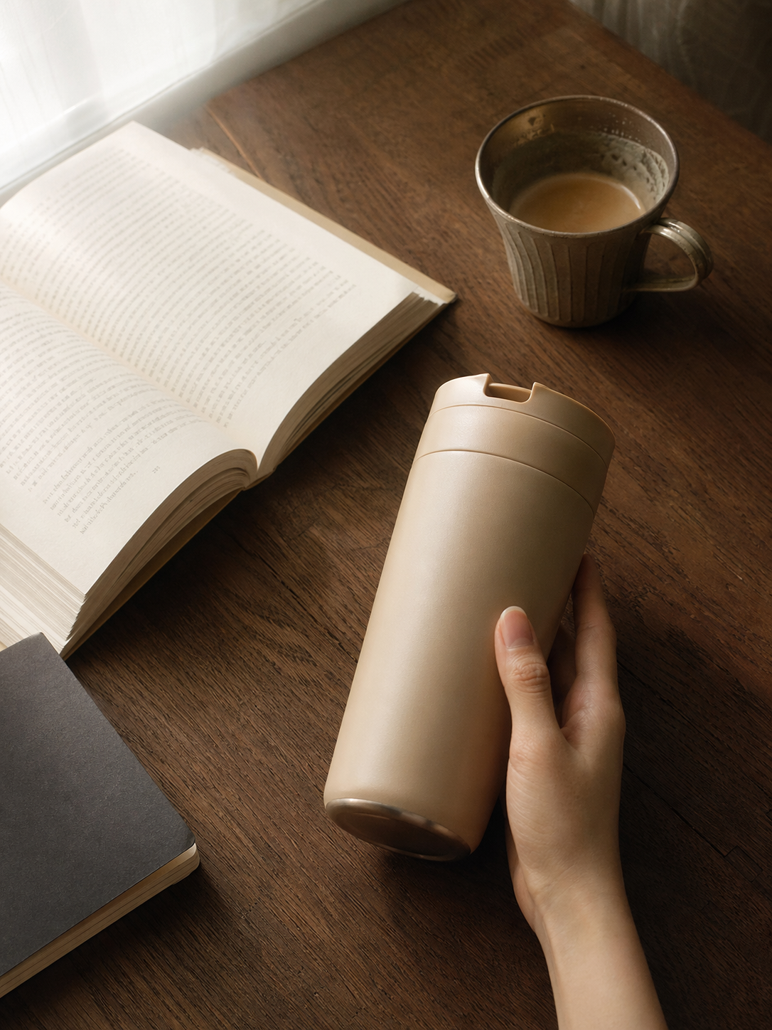 木纹桌面上斜放一只米色保温杯，手部自然扶住杯身一侧，桌边散落笔记本、翻开的书、窗口侧光在杯体留下柔和高光